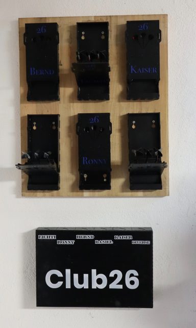 Sechs schwarze Clips auf einer Holzplatte, darunter das Schild „Club26“ für Dartpfeile.