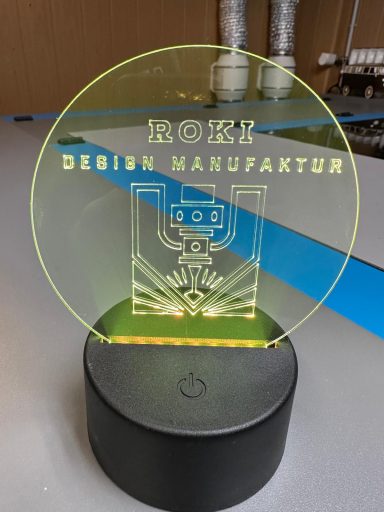 Leuchtendes Acrylschild mit dem Text "ROKI Design Manufaktur" und einem grafischen Design.